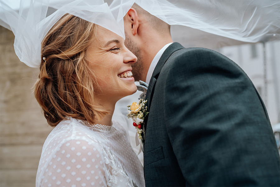 Hochzeit Fotografie, lachende Braut im Profil, dahinter ihr Ehemann, Gesicht halb verdeckt