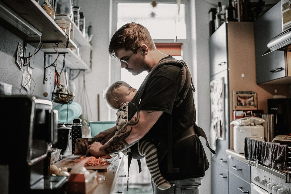 Familienalltag Fotografie, Mann mit Babytrage vor dem Bauch schneidet Möhren in der Küche klein