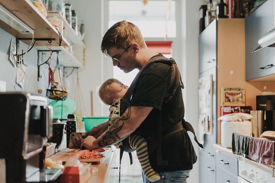Familienalltag Fotografie, Vater in der Küche trägt Kleinkind in Tragetasche vor sich, er schneidet Möhren klein